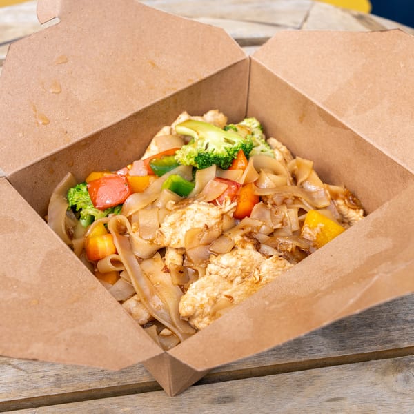 Chicken Pad See Ew with broccoli and bell peppers in a brown takeout box on a wooden table.