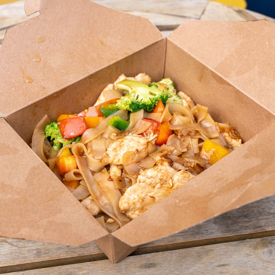 Chicken Pad See Ew with broccoli and bell peppers in a brown takeout box on a wooden table.
