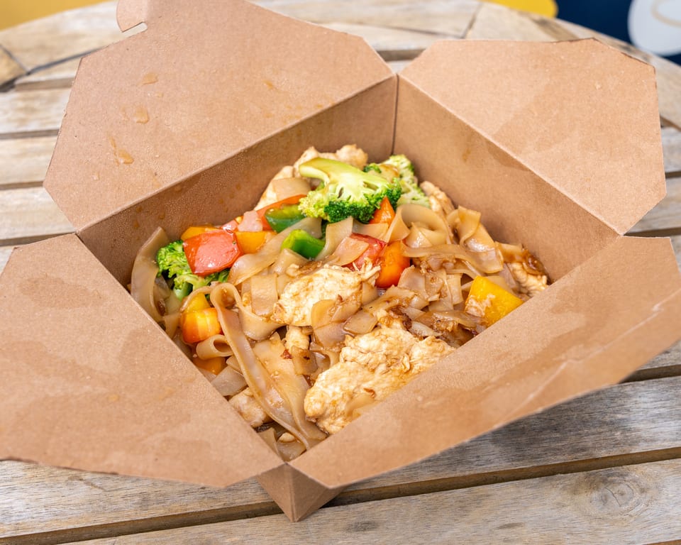 Chicken Pad See Ew with broccoli and bell peppers in a brown takeout box on a wooden table.