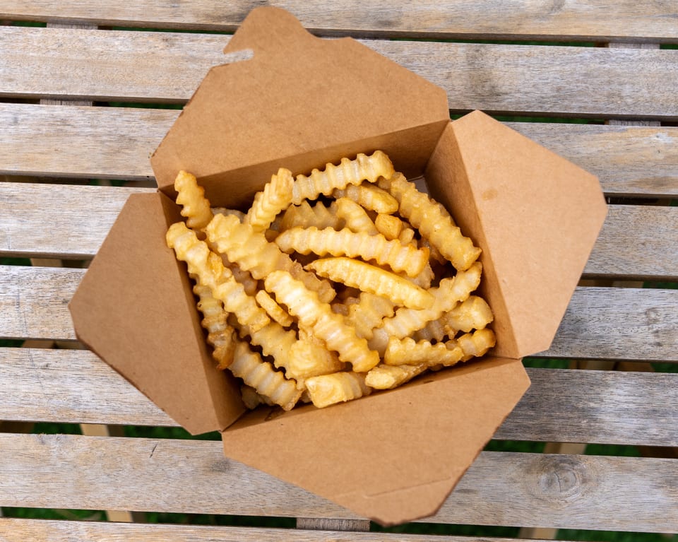 Crinkle-cut french fries in a brown paper take-out container on a wooden slatted table.