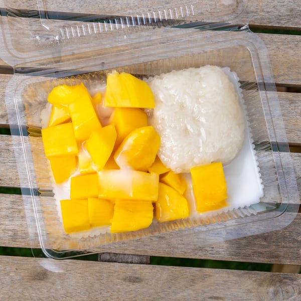 Mango sticky rice with coconut milk in a clear plastic container on a wooden table.