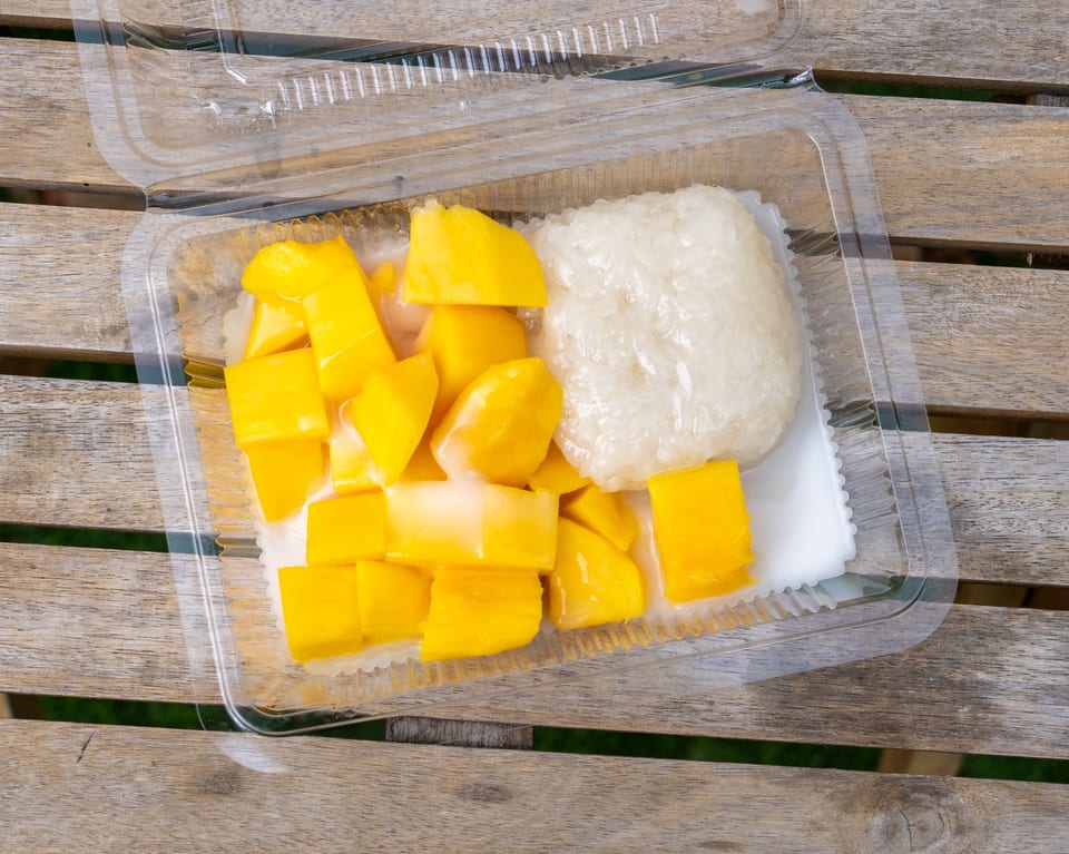 Mango sticky rice with coconut milk in a clear plastic container on a wooden table.