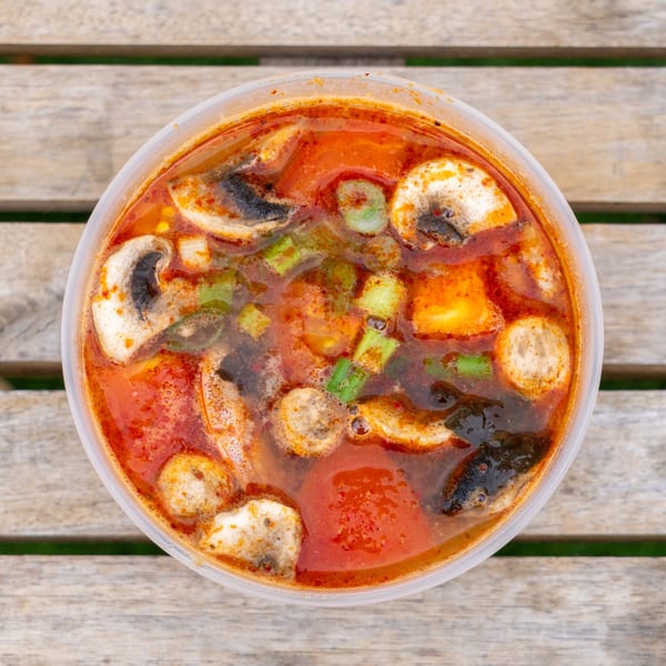 Spicy Tom Yum soup with mushrooms, green onions, and tofu in a clear plastic container on a wooden table.