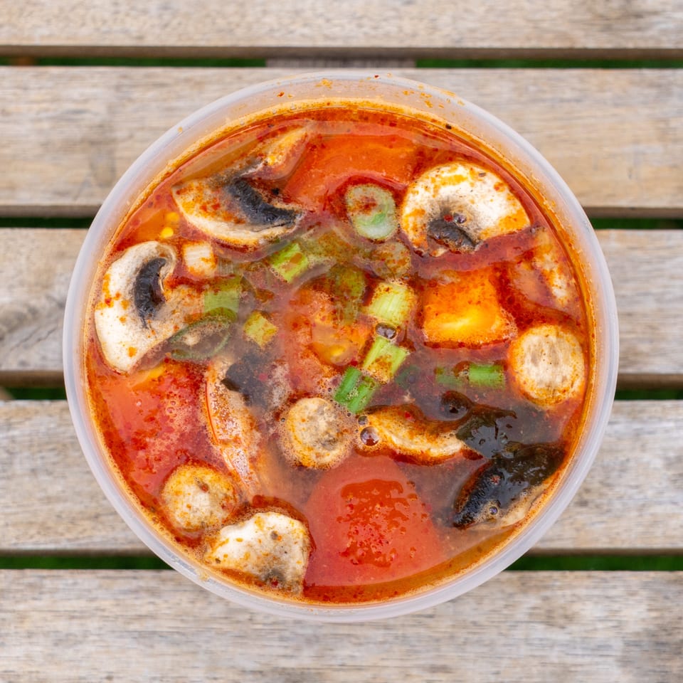 Spicy Tom Yum soup with mushrooms, green onions, and tofu in a clear plastic container on a wooden table.