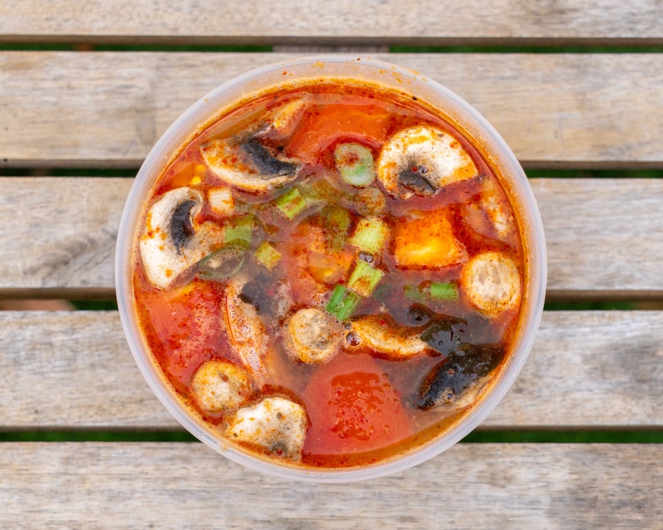 Spicy Tom Yum soup with mushrooms, green onions, and tofu in a clear plastic container on a wooden table.