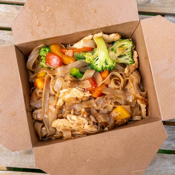 Takeaway box of Pad See Ew with chicken, wide noodles, broccoli, and bell peppers on a wooden table.