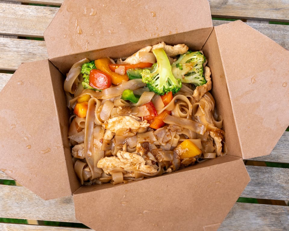 Takeaway box of Pad See Ew with chicken, wide noodles, broccoli, and bell peppers on a wooden table.
