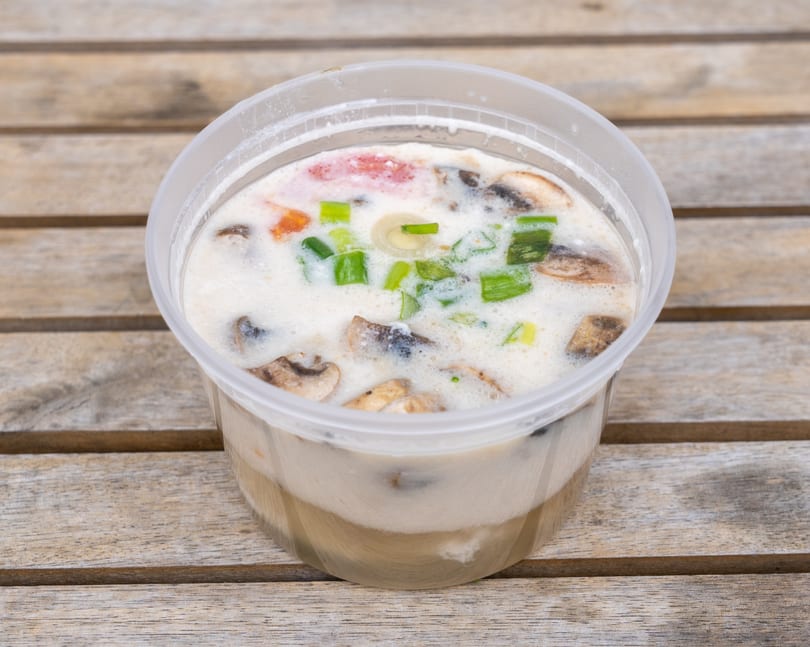 Creamy soup with mushrooms, tomatoes, and green onions in a clear plastic container on a wooden table.