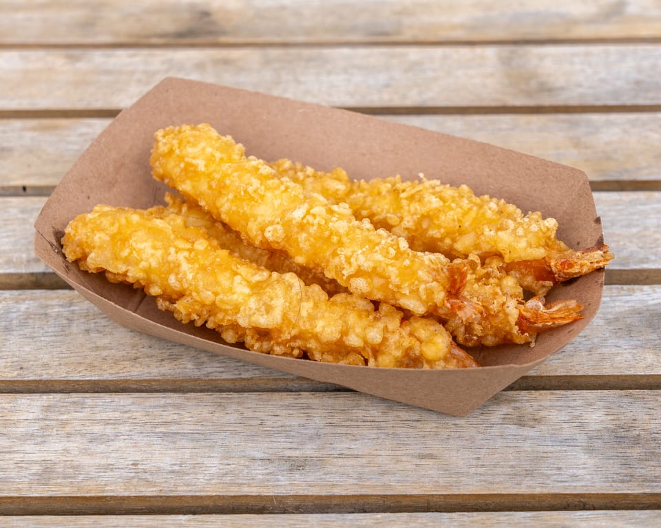 Golden-brown tempura shrimp in a brown paper tray on a rustic wooden table.