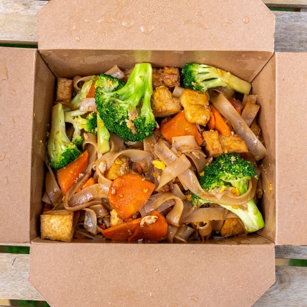 Noodles with broccoli, carrots, and tofu in a brown takeout box on a wooden table.