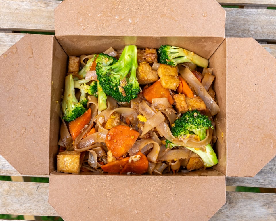 Noodles with broccoli, carrots, and tofu in a brown takeout box on a wooden table.