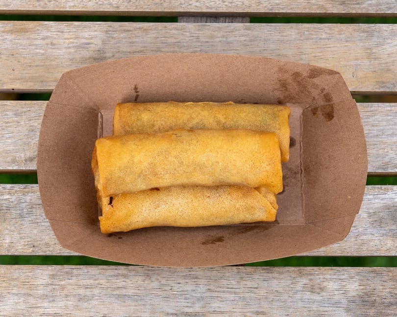 Three golden-brown spring rolls in a brown paper tray, on a wooden slatted table.