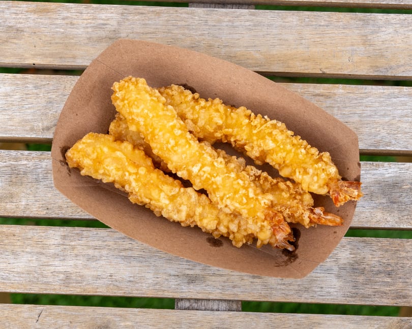 Four golden-brown fried shrimp tempura in a brown paper tray on a wooden picnic table.